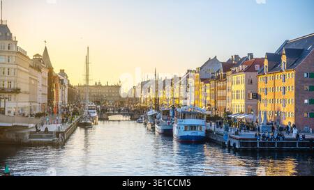 Farbenfrohe Gebäude säumen den Kanal, während die Sonne über Nyhavn in Kopenhagen untergeht. Die Boote schweben sanft auf dem Wasser, während sich die Menschen versammeln, um die lebhafte Atmosphäre zu bewundern. Stockfoto