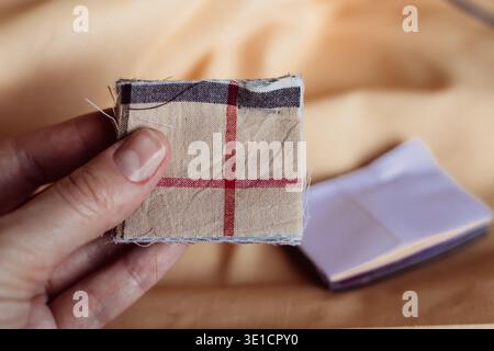 Weibliche Hand, die ein kleines kariertes Stoffmuster über beigefarbenem Textilhintergrund hält. Konzept von Nähhobbys, Modedesigns, Materialauswahl und Babykott Stockfoto