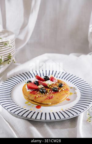 Köstlicher Stapel Pfannkuchen auf Teller mit blauen Streifen, belegt mit Erdbeeren, Heidelbeeren, Pistazien und Schlagsahne, getränkt in Sirup Stockfoto
