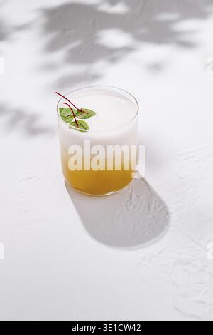 Gelber Cocktail mit weißem Schaum und grünen Blättern wirft Schatten auf weißem Hintergrund Stockfoto