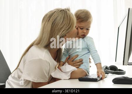 Die junge freischaffende Mutter küsst ihren kleinen Sohn, der auf dem Schreibtisch an ihrem Arbeitsplatz sitzt, auf die Wange Stockfoto