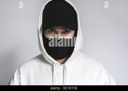 Junger Mann mit weißem Hoodie, schwarzer Baseballmütze und Stoffmaske vor grauem Hintergrund Stockfoto