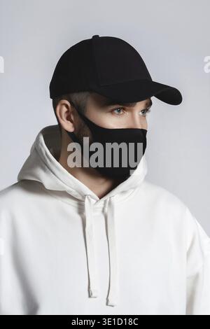 Junger Mann mit weißem Hoodie, schwarzer Baseballmütze und Stoffmaske vor grauem Hintergrund Stockfoto