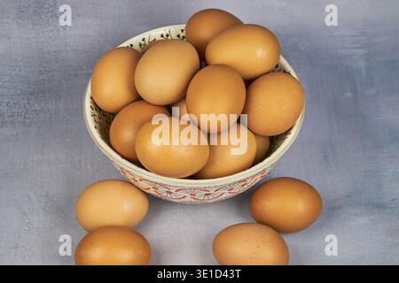 Frische braune Eier in einer dekorativen Schüssel mit mehr Eiern auf einer strukturierten Oberfläche züchten Stockfoto