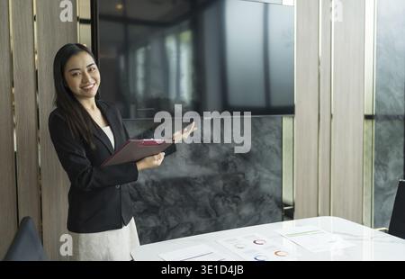 Schöne asiatische Geschäftsfrau in schwarzem Anzug, die auf den Bildschirm zeigt Wall TV, der Digital Entrepreneur zeigt, der ein Projekt auf leerem Geröll präsentiert Stockfoto
