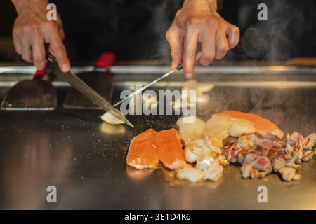 Hände des Küchenchefs mit Spachtel über Teppanyaki. Kochen von Gemüse, Fleisch und Meeresfrüchten auf heißem Hibachi-Grilltisch. Traditionelle Japanische Küche. Teppan Show Stockfoto