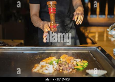 Hände des Küchenchefs mit Spachtel über Teppanyaki. Kochen von Gemüse, Fleisch und Meeresfrüchten auf heißem Hibachi-Grilltisch. Traditionelle Japanische Küche. Teppan Show Stockfoto