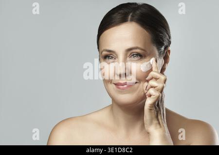 Schöne Frau mittleren Alters Anwendung Anti-Aging-Creme auf ihr Gesicht auf grau Stockfoto