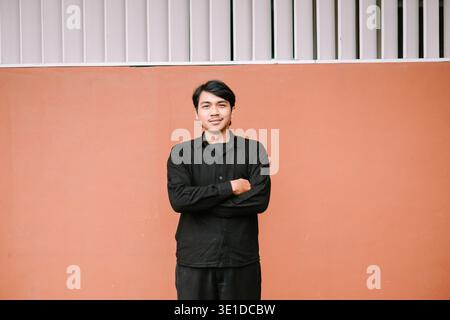 Ein selbstbewusster junger asiatischer Mann steht mit gekreuzten Armen und blickt mit einem freundlichen Lächeln direkt in die Kamera. Er trägt ein schwarzes Hemd gegen einen einfachen pe Stockfoto