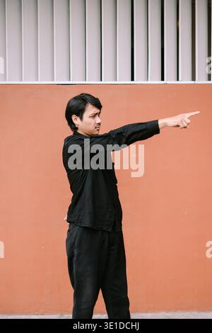 Ein junger Mann in schwarzer Kleidung zeigt mit dem Finger nach rechts und steht vor einer leuchtenden orangen Wand. Sein fokussierter Ausdruck vermittelt Sinn und Regie Stockfoto
