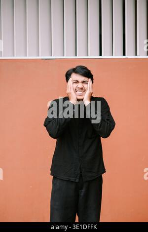 Dieses Porträt fängt einen jungen Mann ein, der seinen Kopf mit einer Grimasse umklammert, was auf Schmerzen, Sorgen oder überwältigenden Stress an einer farbenfrohen Wand hindeutet. Stockfoto