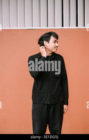 Ein junger Mann in einem schwarzen Langarmhemd steht lässig an einer orangen Wand mit weißen Lüftungsschlitzen und wirkt nachdenklich, während er sich den Kopf kratzt. Stockfoto