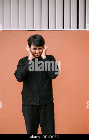 Ein junger Mann mit schmerzhaftem Ausdruck bedeckt seine Ohren, was auf Unwohlsein durch lauten Lärm, Kopfschmerzen oder überwältigenden Stress hindeutet. Dieses Bild stellt den dar Stockfoto