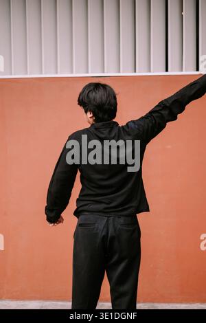 Rückansicht einer stilvollen Person in schwarzer Freizeitkleidung, die draußen mit erhobenem Arm vor einer leuchtend orangefarbenen Wand steht, was auf Freilassung hindeutet Stockfoto