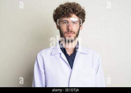 Studio-Aufnahme eines jungen gutaussehenden bärtigen Mannes Doktor mit lockigem Haar als Wissenschaftler, der Schutzbrille vor weißem Hintergrund trägt Stockfoto