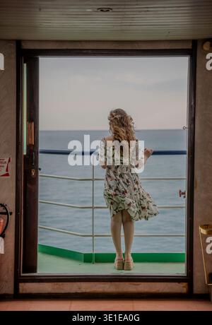 Eine Frau in einem Blumenkleid steht am offenen Geländer eines Fährdecks und blickt auf das ruhige Mittelmeer während einer Überfahrt. Stockfoto