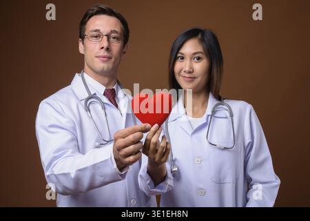 Studio shot Junger stattlicher Mann Arzt und junge schöne asiatische Frau Doktor gemeinsam gegen braunen Hintergrund Stockfoto