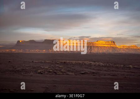 Die Sonne der goldenen Stunde erleuchtet eine riesige mesa in der Wüstenlandschaft, wirft lange Schatten und hebt die zerklüfteten Strukturen der Felsformationen hervor. Stromleitungen erstrecken sich über die Szene, ein Beweis für die menschliche Präsenz in dieser abgelegenen und schönen Umgebung. Stockfoto