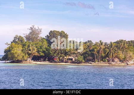 Ein Mann spaziert am Strand einer kleinen tropischen Insel entlang, mit traditionellen Hütten, eingebettet zwischen üppigen grünen Bäumen und Palmen. Das klare blaue Wasser la Stockfoto