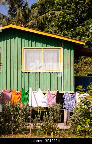 Die Wäsche hängt an einer Wäscheleine vor einem grünen Holzhaus mit gelber Zierleiste. Die Kleidung trocknet in der Sonne, was auf ein warmes Klima hindeutet. Die Stockfoto