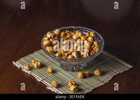 Eine Glasschale hält süßes Popcorn auf einer Bambusmatte. Popcorn ist auf dem Tisch verstreut. Die Einstellung zeigt eine einfache Wohnküche an. Stockfoto