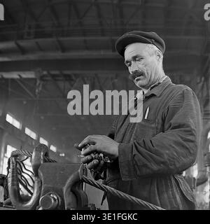 Porträt eines erfahrenen sowjetischen Arbeiters aus den 1960er Jahren mit Schnurrbart, der schwere Stahlkabel in einem Industriewerk in der Region Slawjansk handhabt. Der Mann, der eine klassische flache Kappe und schmutzige Arbeitskleidung trägt, konzentriert sich auf die Sicherung des Metallseils mit einem Industrieknopf. Eine mächtige historische Momentaufnahme, die die physische Arbeit, das Fachwissen und den rauen Charakter der Arbeiterklasse während der Ära der sowjetischen industriellen Expansion hervorhebt. Ein ergreifender Einblick in das Leben vor dem Krieg und den friedlichen Donbass. Stockfoto