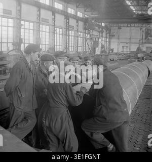 Vintage-Foto aus den 1960er Jahren einer großen Gruppe sowjetischer Fabrikarbeiter, die sich um ein massives Metallrohr versammelten, um technische Baupläne in einer geräumigen Werkstatt in der Region Slawjansk zu überprüfen. Das Team von Männern, die in robuster Arbeitskleidung und flachen Kappen gekleidet sind, führt eine ernsthafte Diskussion über die technischen Pläne. Ein faszinierendes historisches Archivbild, das Teamarbeit, industrielle Problemlösungen und die kollektive Arbeit der sowjetischen Arbeiterklasse erfasst. Ein Blick in das Leben vor dem Krieg und den friedlichen Donbass. Stockfoto