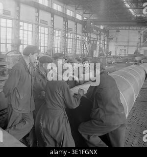 Vintage-Foto aus den 1960er Jahren einer großen Gruppe sowjetischer Fabrikarbeiter, die sich um ein massives Metallrohr versammelten, um technische Baupläne in einer geräumigen Werkstatt in der Region Slawjansk zu überprüfen. Das Team von Männern, die in robuster Arbeitskleidung und flachen Kappen gekleidet sind, führt eine ernsthafte Diskussion über die technischen Pläne. Ein faszinierendes historisches Archivbild, das Teamarbeit, industrielle Problemlösungen und die kollektive Arbeit der sowjetischen Arbeiterklasse erfasst. Ein Blick in das Leben vor dem Krieg und den friedlichen Donbass. Stockfoto