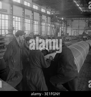 Vintage-Foto aus den 1960er Jahren einer großen Gruppe sowjetischer Fabrikarbeiter, die sich um ein massives Metallrohr versammelten, um technische Baupläne in einer geräumigen Werkstatt in der Region Slawjansk zu überprüfen. Das Team von Männern, die in robuster Arbeitskleidung und flachen Kappen gekleidet sind, führt eine ernsthafte Diskussion über die technischen Pläne. Ein faszinierendes historisches Archivbild, das Teamarbeit, industrielle Problemlösungen und die kollektive Arbeit der sowjetischen Arbeiterklasse erfasst. Ein Blick in das Leben vor dem Krieg und den friedlichen Donbass. Stockfoto