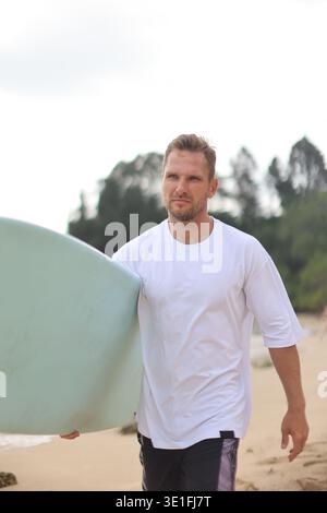 Fokussiertes Mann in weiß T-Shirt in der Nähe des Ozeans. Person Trägt Ein Einfarbiges Weißes Hemd Neben Surfboard By Coast Stockfoto