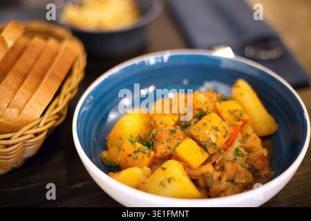 Gelbes Essen auf einer blauen Platte. Gemüseeintopf mit Kartoffeln Karotten Pfeffer mit Kräutern bestreut. Hochwertige Fotonahrung auf einem Holztisch. Die Ansicht von Stockfoto