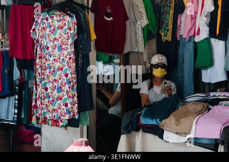 Taiwanesischer Straßenladen mit einem Händler, der eine Maske trägt, umgeben von dicht gehängten bunten Damenkleider, Hemden und Hosen auf Regalen, die an den lokalen Markt erinnern. Stockfoto