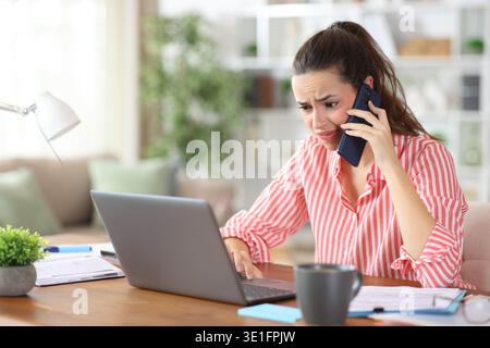Angeekelter Selbstständiger, der einen Laptop überprüft, der am Telefon telefoniert und zu Hause arbeitet Stockfoto