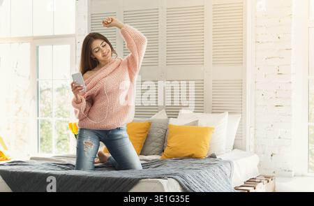 Junge Frau genießt tagsüber Musik, während sie auf dem Bett sitzt, in einem hellen Zimmer mit großen Fenstern Stockfoto