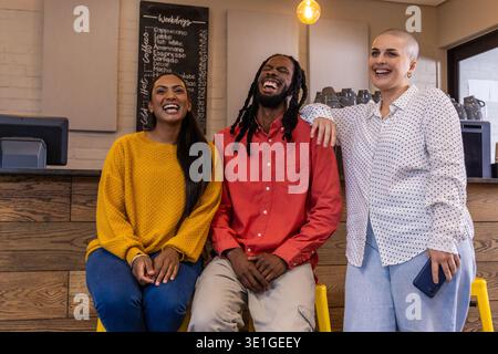 Verschiedene Freunde sitzen am Coffee Shop-Theke auf gelben Hockern, lachen und schauen nach dem Smartphone Stockfoto