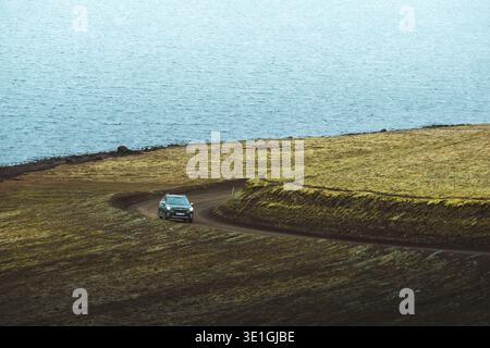 Blick auf ein Auto, das auf einer gewundenen Feldstraße durch die goldene isländische Landschaft vor dem Hintergrund des ruhigen blauen Meeres, Rangárþing ytra, IC, fährt Stockfoto