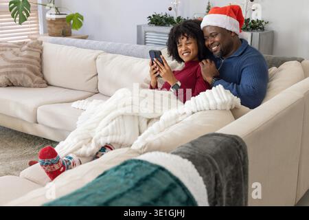 Afroamerikanisches Paar kuschelt sich zu Hause unter Decke auf Sofa, trägt Weihnachtsmütze und hält Telefon. Gemütlich, Zuneigung, Strick, Couch, Smartwatch, garland, festlich || Model veröffentlicht Stockfoto