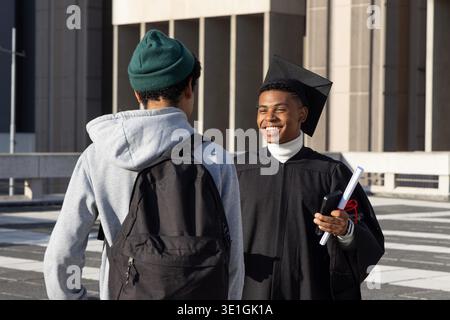 Verschiedene Männer gratulieren zum Abschluss in Mütze und Kleid mit Diplom und Telefon auf dem Campus plaza. Outdoor, Architektur, Säulen, Beton, Rucksack, beanie, Hoodie || Model veröffentlicht Stockfoto