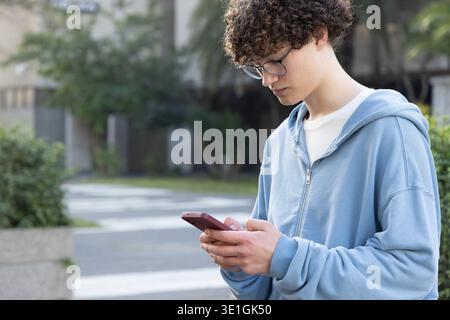 Männlicher Teenager, der auf dem Campus steht, einen blauen Hoodie trägt, eine Brille mit Smartphone, einen Kopierraum. Innenhof, Fußgängerübergang, Pflanzgefäße, Grün, Bäume, diffused, Technologie || Modell veröffentlicht Stockfoto