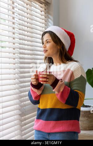 Frau, die zu Hause in der Nähe von Jalousien steht und einen roten Becher mit gestreiftem Pullover und Weihnachtsmann-Hut trägt. Fenster, Tageslicht, innen, gemütlich, besinnlich, festlich, Kopfbedeckung || Model veröffentlicht Stockfoto