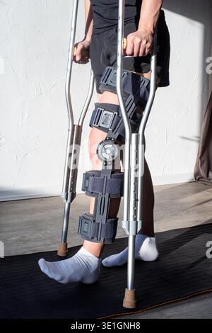 Frau nach Autounfall in einer Orthese, die auf einer Sportmatte steht. Frau mit Knieorthese oder Kniestützstrebe und Krücken nach Beinoperationen Stockfoto