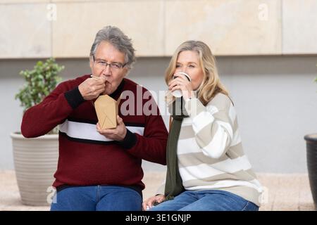 Seniorenpaar sitzt auf einer Bank im Innenhof und isst aus dem Take-Out-Container und schlürft Kaffee. Älter, Begleiter, Outdoor, plaza, Stadtbild, lässig, entspannt || Model veröffentlicht Stockfoto