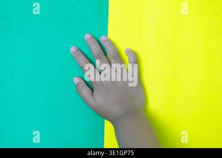 Eine kleine Hand wird sanft auf einen hellen Hintergrund gelegt, der gleichmäßig zwischen leuchtendem Grün und sonnigem Gelb aufgeteilt wird. Diese farbenfrohe, minimalistische Komposition Stockfoto