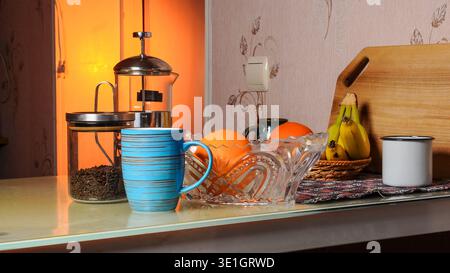 Eine Stillleben-Komposition mit einer französischen Presse gefüllt mit Kaffeebohnen, einem blauen Becher, einer Kristallschale mit Orangen, Bananen in einem Korb und einem weißen Becher Stockfoto