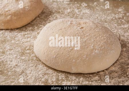 Zwei Runden rohen Pizzateigs, bedeckt mit einem leichten Staubbeutel Mehl, stehen auf einer bemehlten Oberfläche, bereit zum Backen Stockfoto
