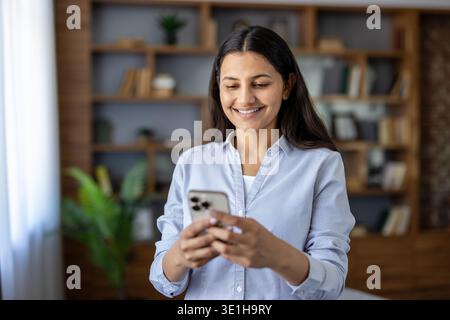 Glückliche junge indische Frau, die drinnen steht, lächelt, während sie auf einem Smartphone SMS schreibt, soziale Medien und Online-Verbindung in entspannter Atmosphäre zu Hause oder im zwanglosen Büro genießt Stockfoto