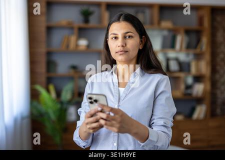 Junge Frau, die in einem Heimbüro steht und ein Smartphone hält, repräsentiert moderne Kommunikation, Online-Arbeit und digitale Technologie an einem entfernten Arbeitsplatz Stockfoto