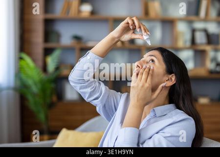 Junge Frau, die Augentropfen aufträgt, während sie zu Hause auf einem Sofa sitzt, um gereizte Augen zu lindern, Allergien zu behandeln oder das Syndrom des trockenen Auges zu bekämpfen Stockfoto