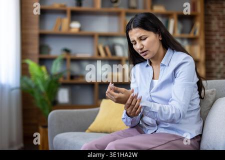 Junge Frau auf einem Sofa massiert ihr Handgelenk und Hand mit Schmerzen, zeigt Beschwerden durch Arthritis, Karpaltunnel oder Verletzung zu Hause, müde und frustriert Stockfoto