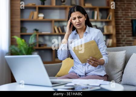 Eine gestresste junge Frau, die unerwartet schlechte Nachrichten in einem Brief erhält, leidet unter Kopfschmerzen, während sie zu Hause auf einem Sofa sitzt, mit einem Laptop und Dokumenten auf dem Tisch Stockfoto